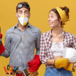bug-eyed-male-wearing-protective-mask-gloves-having-tool-belt-waist-holding-drill-being-scared-difficulties-much-work-standing-near-his-colleague-who-is-looking-him-with-smile_273609-7928