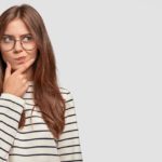 confused-lovely-female-teenager-holds-chin-looks-thoughtfully-aside-has-dark-hair-wears-striped-sweater-isolated-white-wall_273609-16546