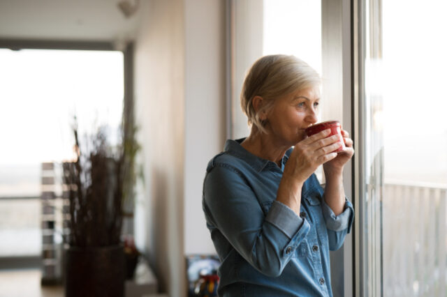 beautiful-senior-woman-at-home-standing-at-the-window-in-her-living-room-holding-a-cup-SBI-305234432 Asset ID: SBI-305234432