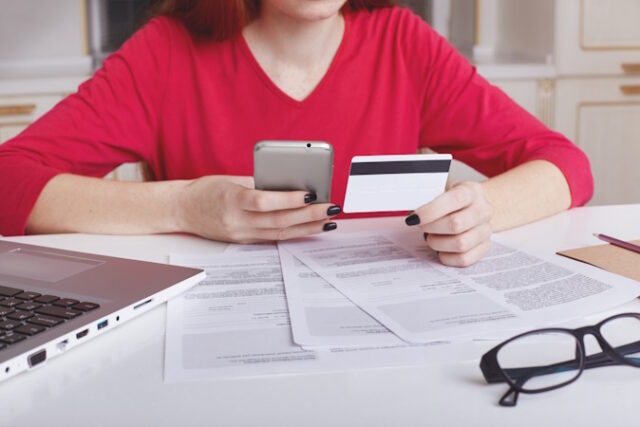 Cropped shot of unrecognizable female model in red sweater sits at working table surrounded with papers and laptop computer, uses cell phone for paying bill online with credit card. Ecommerce concept https://www.freepik.com/free-photo/unrecognizable-female-model-red-sweater-sits-working-table-surrounded-with-papers-laptop-computer_8988194.htm#query=credit%20check&position=34&from_view=search&track=ais