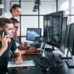 Guy talks by phone. Team of stockbrokers works in modern office with many display screens