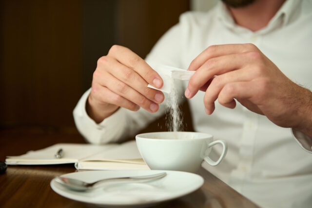 Close-up of an unrecognizable man in a white shirt sprinkling sugar from sticks into a white ceramic cup with a freshly brewed coffee drink, sitting at a wooden table and enjoying a coffee break https://www.vecteezy.com/photo/27031225-close-up-of-an-unrecognizable-man-in-a-white-shirt-sprinkling-sugar-from-sticks-into-a-white-ceramic-cup-with-a-freshly-brewed-coffee-drink-sitting-at-a-wooden-table-and-enjoying-a-coffee-break
