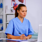 Medical assistant typing on laptop and taking notes on clipboard