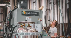 A woman controls an Earthly Labs-branded machine in a factory setting; a tank that says “carbon dioxide” is in the foreground.