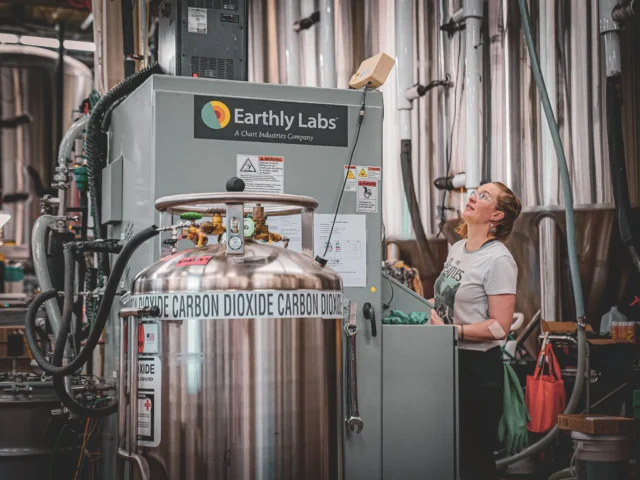 A woman controls an Earthly Labs-branded machine in a factory setting; a tank that says “carbon dioxide” is in the foreground.