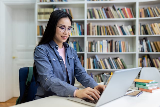 Asian woman studies in library, working on a laptop with books on the shelves. https://www.vecteezy.com/photo/68905002-a-woman-with-glasses-using-a-laptop-in-a-library-with-bookshelves-in-the-background-she-is-studying